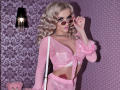 MademoiselleBella with blonde curls, wearing pink sheer lingerie, pink fur-trimmed jacket, pink sunglasses, holding white strap, necklace, tattoo on right side, purple-patterned wall, black lamp.