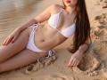 Rouzi, con cabello largo y castaño, vestida con un bikini blanco, acostada en una playa arenosa, mostrando sus pequeños pechos y glúteos, uñas rojas, manos en la arena, piernas ligeramente separadas, cabeza inclinada..