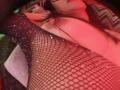 ZarayFeet's large tits are covered in black fishnet with sparkling sequins, her arm is raised, wearing a chevron-patterned garment, red lighting, and partially visible face.