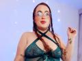 StacyMillerr with curly brown hair, glasses, and green lace lingerie, showing her tits and a small tattoo on her right wrist, smiling confidently.