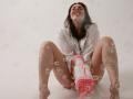 Dreia, with long dark hair, sits on a white floor, legs spread, wearing an open white shirt, smiling, holding a pink and white electric toothbrush, with bubbles around.