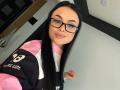 Olieri with long black hair, glasses, and pink-black-white sports jersey, showing a subtle smile, her tits barely visible under the jersey.