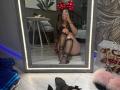 ElwandaKiehne in black lingerie with sheer thigh-high stockings and red polka-dot bow headband, sitting in front of a mirror, showing her ass and tits, holding a phone.