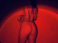 Tisia stands in red lingerie, showcasing her ass and back. She's wearing a thong, her hands near her waist. Her long hair is down, partially visible.