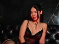MelodyGloss with curly black hair, wearing a sheer black dress, small tits, and fishnet stockings, sitting on a black tufted leather couch, smiling at the camera.