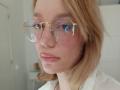 MissFrenchy with shiny pink lips, clear round glasses reflecting light, and shoulder-length blonde hair, wearing a white shirt, looking serious.