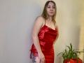 Aminas with long blonde hair, wearing a red satin dress, standing, hands on hips, showing a tattoo on her right thigh, and a potted plant to the right.