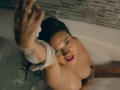 LindaJimenex in a bathtub, reaching up with soapy hands, looking at the camera with red lips, naked, wet brown hair, tattoo on right arm, smiling.