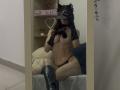 Valnes with cat ears, black lace gloves, black thong, and black knee boots, showing her tits, sitting on a bed, taking a mirror selfie, holding a phone.
