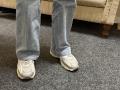 Dalaila's legs in blue jeans with white, slightly dirty running shoes standing on gray carpet. Visible from knees down, wearing worn-out sneakers, showing some dirt.