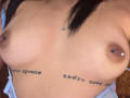 Close-up of EmmaWaltter's chest showing medium-sized, natural breasts with dark areolas and small, blue nipple piercings. Her skin is light brown, and she has black hair. Tattoos with black text are on each side of her breasts. The image has a realistic, unfiltered style with clear details of skin texture and piercings. EmmaWaltter's torso is partially visible, with a light blue garment hanging off her shoulders.