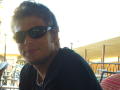 FrancimooR with black sunglasses, short brown hair, and a black shirt, sporting a slight smile, has a light beard and a white shirt underneath.