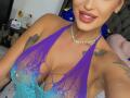 NicolSNaya with tan skin, red lipstick, and a sexy smile, wearing a purple and blue lace-up halter top that reveals her big tits, and a diamond necklace.