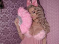 MademoiselleBella with blonde curls, pink bow, and fluffy pink dress, holding a pink fan, showing her big boobs, wearing pink lipstick and a choker, in a pink-themed setting.