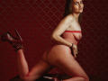 RubiAnderson with red fishnet bodysuit, showing her ass, in high heels, on a leather surface, kneeling, looking at the camera with a slight smile.