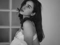 PeacHub69 has long dark hair, staring with serious eyes, wearing a white bra, looking over shoulder with slightly parted lips, soft grayscale, holding a pillow.