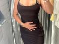 MargoFun with tan skin, wearing a tight black dress, hand on her ass, brown bra visible, silver bracelet, and a watch, standing in a bathroom.