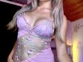 EliseMonet portant une robe rose, transparente, brodée de fleurs, laissant entrevoir ses seins dans un haut à décolleté bas et sans couture à l'entrejambe, avec de longs cheveux blonds et un collier en or..