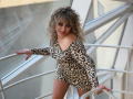 CandyLadyZ with curly blonde hair, wearing a leopard print dress, leaning on a staircase, showing her bare shoulder and smiling with pink lipstick. Her tits are visible.