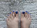 Close-up of HeartlessQueenn's feet with blue-painted toenails, standing on a textured, white carpet. The toes are slightly spread apart, showing some tension in the skin and nails.