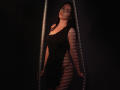 TriciaBarnes with long dark hair, wearing a black dress, standing in a shadowed, striped light environment, holding metallic bars, showing her bare ass and small, perky tits.