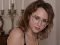 TeresaBirt with wavy brown hair, glasses, and a cute face, wearing a black spaghetti strap top, showing off her tits in a soft-lit room.