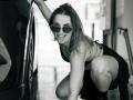 Milagres crouches by a car, tongue out, wearing sunglasses, a black bikini top, and skirt, with wet hair and a playful grin. Her ass is partially visible.