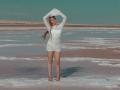 StellaLane stands barefoot in a white lace dress, arms raised, long brown hair flowing. She's got a curvy ass and a cute, confident look. Desert landscape in background.