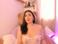 ElyRoss with dark hair, pink lipstick, hoop earrings, and sheer white lingerie showing her tits, sitting on a pink bed with a large teddy bear. Soft pink lighting.