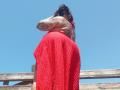 Chili, topless, with tattoos, wearing a red, textured skirt, standing against a clear blue sky, hands in black hair, wooden fence in background, visible ass.