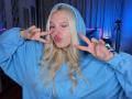 AliceWhitey, blonde, winking, pouting, wearing a blue hoodie, making peace signs with fingers, long wavy hair, blue-green nails, hood up, cute, indoor, soft lighting, casual.