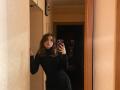 LeScharwath with wavy brown hair, black turtleneck dress, standing in hallway, taking mirror selfie, right hand on wall, holding phone with pink patterned case, looking slightly bored.