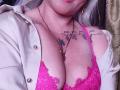 AlessadraJoness with pink lace bra, showing big tits, white shirt open, tattoos on chest, necklace, and silver earrings, pink lips, pale skin, and white hair.