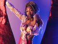 DaniaMarcell with braided hair, wearing a sheer pink top with reflective nipples, pointing forward with a confident expression, red and blue lighting, exposing her tits.
