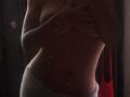 GabrielaD's tits are covered by her hand, showing a slim, dark-skinned torso with grey pants, in a dimly lit, grainy photo.