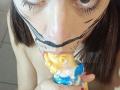 OrianaLaFrancaise, avec des marques noires peintes sur son visage, des yeux marron et des ongles rouges, suce une glace jaune et bleue, la langue tirée vers l'extérieur, et la tient avec sa main droite..