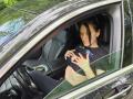 JoliDanielle, with black hair, sits in a car, wearing a black top, touching her small tits, looking down, hands on her chest, in a gray car, green outside.
