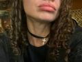 HotCaramelsTrio with curly brown hair, nose piercing, pinkish lips, black choker with cross necklace, wearing black shirt, slightly visible black jacket, medium skin tone, subtle neck muscle.