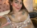 Catalina Reiyna, con un sorriso, capelli castani e un leggero trucco rosa, indossa una camicetta trasparente con un motivo a margherite, che lascia intravedere il seno e i tatuaggi sul petto. Sta scattando un selfie..