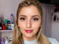 EliseMonet has blonde hair, brown eyes, and pink lipstick, with long eyelashes and pearl earrings, looking directly at the camera with a slight smile. She's wearing a white shirt.