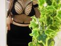 Trima with big tits in black fishnet top, black lace bra, and black skirt, standing next to green and yellow leafy plant. Her hand's in her skirt, nails painted red.