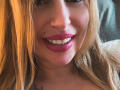 XimenaBella's face, showing a smile with red lipstick, a nose piercing, and blonde hair, with a tattoo on her neck and a white shirt visible.