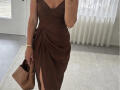 Sloaneswan porte une robe marron avec un décolleté en V plongeant et une fente haute, elle tient un sac beige et porte un bracelet doré dans une pièce lumineuse..