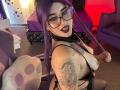 LilithDark69 with purple hair, glasses, black lipstick, and tattoos, showing off her tits in a black latex outfit, tongue out, in a bedroom with purple furniture.