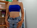 Steisii with a blue strapless top, exposing her tits, and unbuttoned, ripped jeans, standing with hands on her ass, showing a tattoo on her right arm.