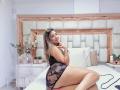 KendraMiler with long blonde hair, wearing black lace lingerie, sits on a bed, showing her ass, looking back with a finger to her mouth, smiling.