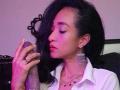 AmelieMartinezO with black hair, pink eyeshadow, and white shirt, kissing a large, dark, erect penis while wearing a diamond necklace and earrings. Her hand has a ring.