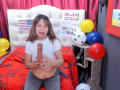 SofiaXDelRio, with brown hair, in a white top, holding a large, erect, circumcised penis, sitting on a red bed with balloons and soccer ball in a colorful room.