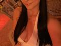 Olieri with long black hair, smiling, wearing a white, deep cleavage, sleeveless top, showing her tits. Warm, red-lit, indoor setting. Soft, relaxed, casual vibe.