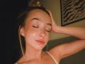 DanikaPlassmann with eyes closed, blonde hair in a bun, hand on head, shiny skin, pink lips, white strap, subtle makeup, small earrings, soft smile, dim lighting.